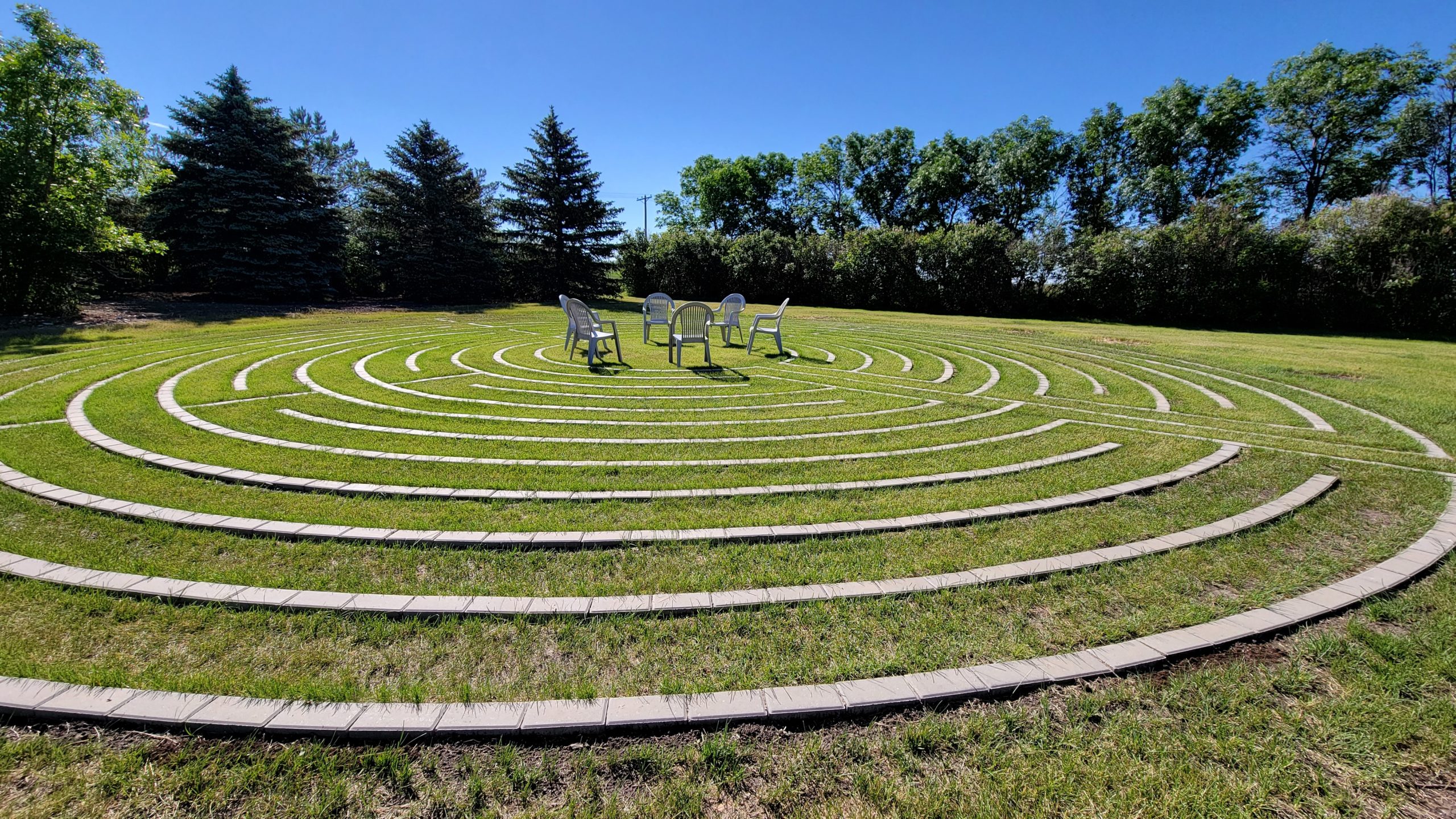 a labrynth or maze laid out with bricks in the grass and surrounded by trees. at the centre are a circular cluster of chairs facing each other.