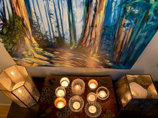 A tray of candles lit in front of a sureal blue painting of rainforest trees.